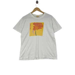 May include: White t-shirt with a graphic print of a red and orange flower on a yellow background. The text "The Flower" is printed below the flower.