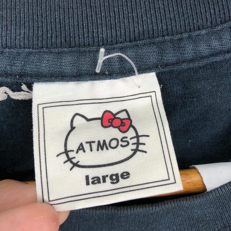 May include: Close-up of a white tag with a black border, featuring a cartoon cat head with a red bow and the word "ATMOS" in black. Below, the word "large" is printed. The tag is attached to a dark blue t-shirt.