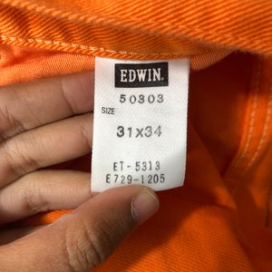 Op de afbeelding: Een wit label met zwarte tekst dat "EDWIN."  "50303"  "SIZE 31x34"  "ET-5313 E729-1205" aangeeft.