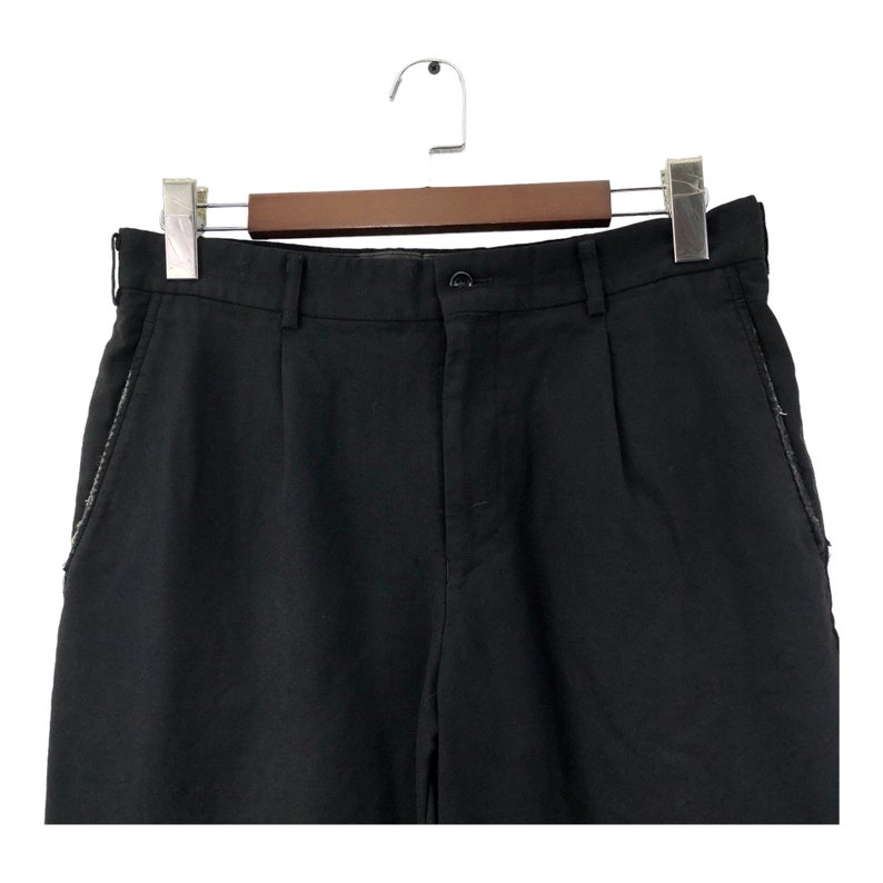 May include: Black pants with a button closure and belt loops, hanging on a wooden hanger. The pants have a classic design with a straight leg and a tailored fit. The pockets have a decorative trim.