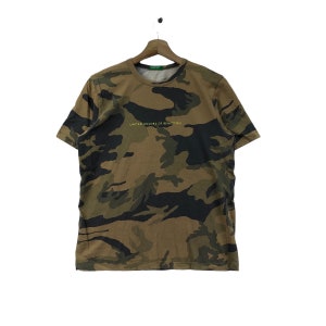 Puede incluir: Una camiseta de manga corta y cuello redondo con estampado de camuflaje. La camiseta tiene el texto "UNITED COLORS OF BENETTON" impreso en amarillo en la parte delantera.