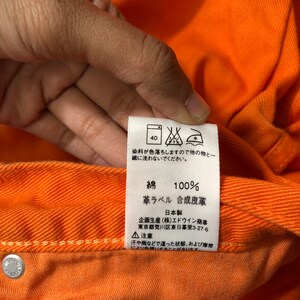 Op de afbeelding: Een close-up van een feloranje stoffen label met wasinstructies. Het label zegt "Kleurstof kan afgeven, dus apart wassen van andere items." Het label zegt ook "Katoen 100%", "Lederen label Kunstleer", "Gemaakt in Japan", "Gepland en geproduceerd door Edwin Trading Co., Ltd., Tokio, Japan".