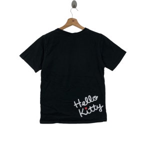 K&ouml;nnte beinhalten: Schwarzes T-Shirt mit kurzen &Auml;rmeln und einem wei&szlig;en "Hello Kitty"-Motiv unten rechts. Das Motiv enth&auml;lt ein kleines rotes Herz.