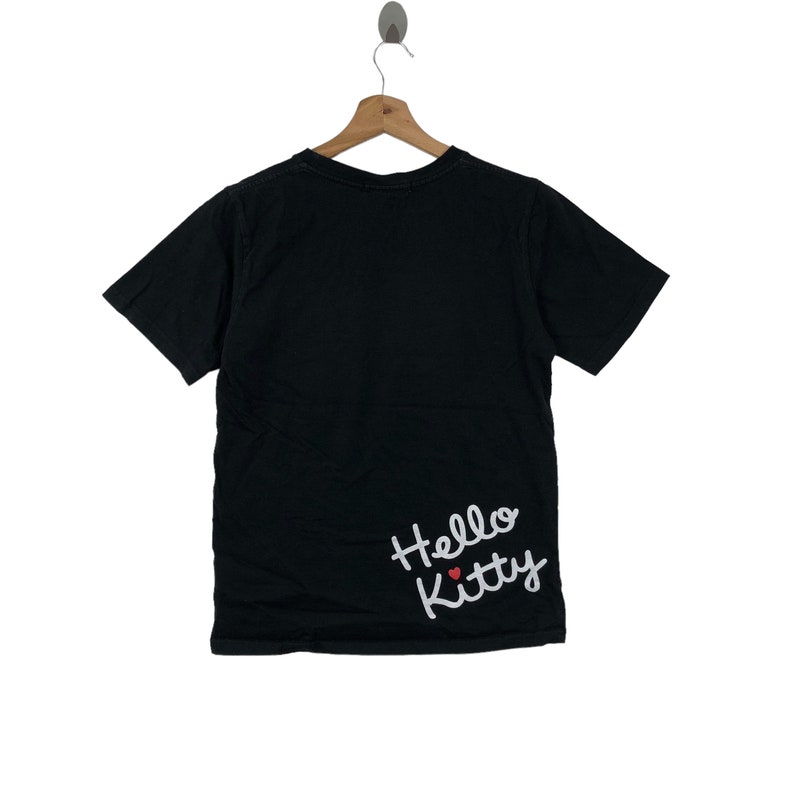 K&ouml;nnte beinhalten: Schwarzes T-Shirt mit kurzen &Auml;rmeln und einem wei&szlig;en "Hello Kitty"-Motiv unten rechts. Das Motiv enth&auml;lt ein kleines rotes Herz.