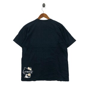 May include: Black t-shirt with a graphic of a cartoon cat with a bow and the word "ATMOS" in white. The t-shirt is hanging on a wooden hanger against a white background.