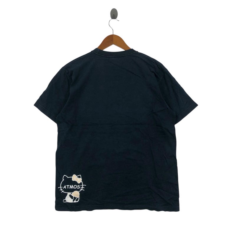 May include: Black t-shirt with a graphic of a cartoon cat with a bow and the word "ATMOS" in white. The t-shirt is hanging on a wooden hanger against a white background.