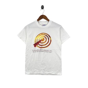 May include: White t-shirt with a graphic of a target with a red arrow and concentric circles in shades of yellow and brown. The text "CharisMac Engineering, INC." and a tagline are printed below the graphic.