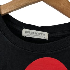 K&ouml;nnte beinhalten: Schwarzes T-Shirt mit einem gro&szlig;en roten Kreis auf der Vorderseite. Das Shirt hat ein wei&szlig;es Etikett mit dem Text "HELLO KITTY &copy;1976, 2016 SANRIO CO., LTD. TOKYO, JAPAN &copy;"