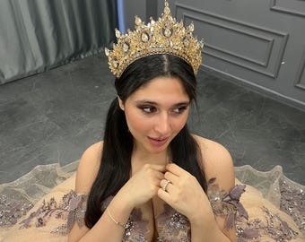 Puede incluir: Una corona dorada con diamantes de imitación transparentes, puesta por una persona con cabello largo y oscuro. La corona tiene un diseño puntiagudo con una piedra central grande.