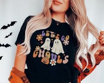 Könnte beinhalten: Ein schwarzes Kurzarm-T-Shirt mit einem Grafikdruck, der den Text "LET'S GO GHOULS" in einer Retro-Schriftart zeigt. Das Design umfasst zwei weiße Geister, einen kleinen orangefarbenen Kürbis und mehrere bunte Blumen in Orange-, Rosa- und Violetttönen.