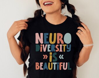Puede incluir: Una camiseta negra de manga corta con el texto multicolor NEURO DIVERSITY IS BEAUTIFUL en una fuente redondeada y negrita. Las letras son de color rosa, amarillo, verde azulado y azul claro, con pequeños símbolos de chevrón flanqueando la palabra IS.