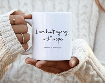 Könnte beinhalten: Weiße Keramik-Tasse mit dem Zitat "I am half agony, half hope" aus Jane Austens Persuasion. Die Tasse wird von einer Person gehalten, die einen weißen Pullover trägt. Die Tasse hat eine Standardgröße und -form.
