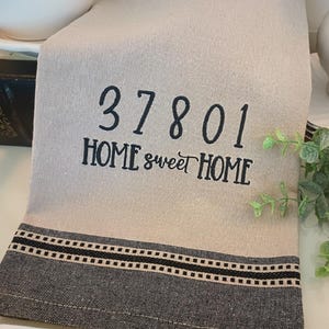 May include: A beige kitchen towel with black embroidered text that reads "37801 HOME sweet HOME". The towel has a black and white striped border.