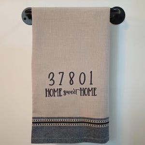 Peut inclure: Une serviette de cuisine beige avec un motif brod&eacute; noir qui dit "37801 HOME sweet HOME". La serviette a une bordure ray&eacute;e bleue et noire.