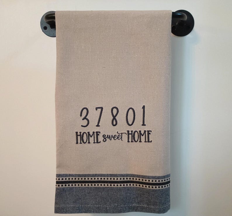 Peut inclure: Une serviette de cuisine beige avec un motif brod&eacute; noir qui dit "37801 HOME sweet HOME". La serviette a une bordure ray&eacute;e bleue et noire.