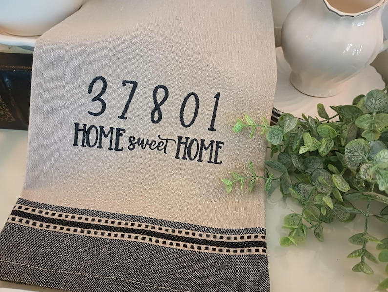 Peut inclure: Une serviette de cuisine beige avec un texte brod&eacute; noir qui dit "37801 HOME sweet HOME". La serviette a une bordure ray&eacute;e noire et blanche.