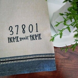 Peut inclure: Une serviette de cuisine beige avec une bordure brod&eacute;e noire et le texte "37801 HOME sweet HOME".