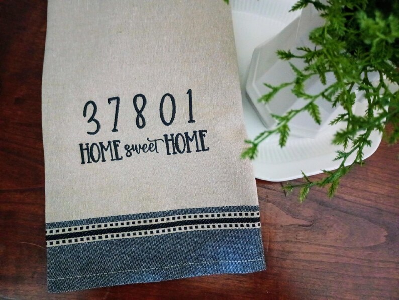 Peut inclure: Une serviette de cuisine beige avec une bordure brod&eacute;e noire et le texte "37801 HOME sweet HOME".