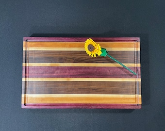 May include: A rectangular wooden cutting board with a natural wood finish. The board has a decorative edge and is made of multiple wood types, including purpleheart, walnut, and maple. A small yellow sunflower is placed on the board.