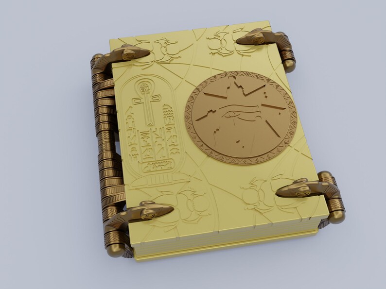 May include: A gold-colored book with intricate designs and a circular emblem featuring an eye and a symbol. The book has two clasps on each side.