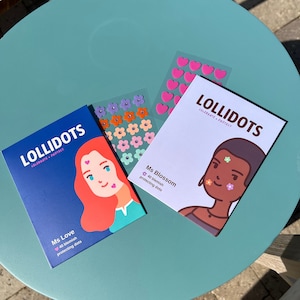 May include: Three packages of LOLIDOTS blemish-protecting dots are displayed on a teal table. The packages are blue and white, with illustrations of faces and the text "Ms Love" and "Ms Blossom." Two sheets of decorative stickers with pink hearts and colorful flowers are also visible.