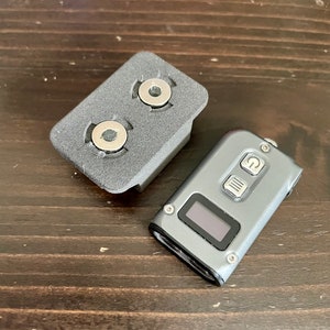 May include: A grey metal device with a digital display and three buttons. The device is sitting on a wooden surface next to a grey plastic base with two circular cutouts.