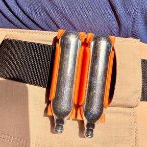 May include: Two silver CO2 cartridges in an orange holder attached to a tan belt. The belt has a black woven strap. The cartridges are held against tan trousers and a blue shirt.