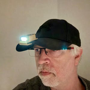 May include: A person wearing a black baseball cap with a headlamp attached to the front. The headlamp is on and illuminating the person's face. The person is also wearing glasses.