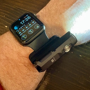 Op de afbeelding: Een zwarte smartwatch met een digitaal klokdisplay dat de tijd 17:26 en de datum DO 21 weergeeft. Het horloge is bevestigd aan een zwarte polsband met een zilveren zaklamp die aan de band is bevestigd.
