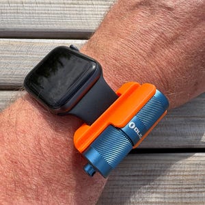 Könnte beinhalten: Eine schwarze Smartwatch mit schwarzem Armband ist an einem Handgelenk befestigt. Eine orangefarbene und blaue Taschenlampe ist am Armband befestigt. Auf der Taschenlampe steht der Markenname "OLIGHT".