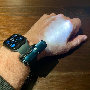 Może przedstawiać: Czarny Apple Watch z niebieską latarką przymocowaną do paska. Tarcza zegarka wyświetla godzinę 10:19:16.