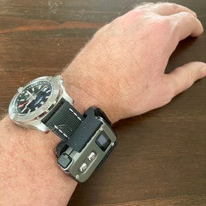 Op de afbeelding: Een zilveren polshorloge met een zwarte wijzerplaat en een zwarte nylon band. Een klein zwart en grijs apparaat is aan de band bevestigd.