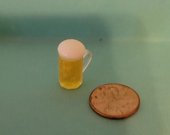 May include: A miniature glass beer stein with a white foam head and a yellow liquid inside. The stein is next to a US penny.