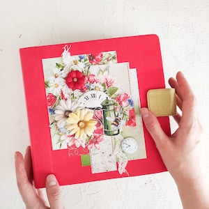 May include: Red journal with a floral design on the front cover. The design includes a teapot, a bee, and a clock face. The journal has a gold clasp.
