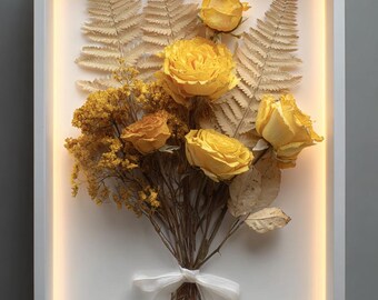 May include: A framed floral arrangement featuring yellow roses, dried ferns, and other dried flowers. The bouquet is tied with a white ribbon. The frame has a white border with a warm, glowing light.