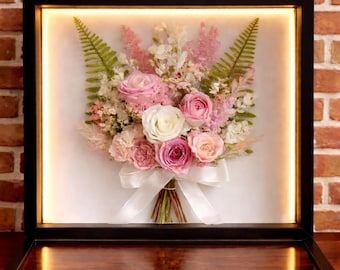 May include: A square black shadow box frame containing a preserved floral arrangement of pink and white roses, ferns, and small filler flowers. The bouquet is tied with a white satin ribbon and illuminated by an integrated LED light strip.