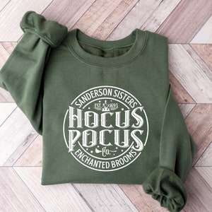 May include: A green sweatshirt with a white circle graphic that says "Sanderson Sisters Est. 1693 Hocus Pocus Co. Enchanted Brooms"