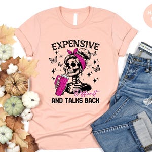 May include: A pink t-shirt with a black and white graphic of a skeleton with a pink bow in her hair. The text on the shirt reads "Expensive, Difficult and Talks Back".