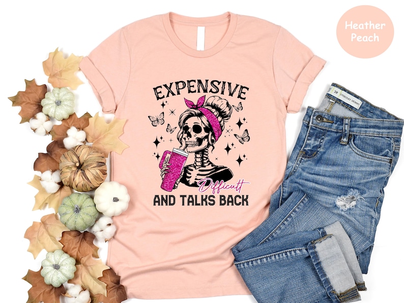 May include: A pink t-shirt with a black and white graphic of a skeleton with a pink bow in her hair. The text on the shirt reads "Expensive, Difficult and Talks Back".