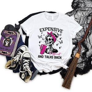 May include: White t-shirt with a black and white illustration of a skeleton woman with a pink bow in her hair. The skeleton is holding a pink cup and the text "Expensive and Talks Back" is printed below the illustration.