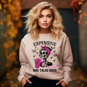 May include: A light brown sweatshirt with a graphic of a skeleton wearing a pink bandana and holding a pink cup. The text on the sweatshirt reads "Expensive, Difficult and Talks Back".