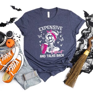 May include: A navy blue t-shirt with a white graphic of a skeleton with a pink bow holding a pink drink. The text "Expensive, Difficult and Talks Back" is printed below the skeleton.