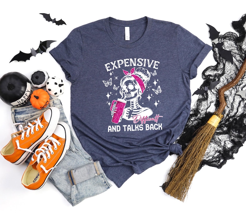 May include: A navy blue t-shirt with a white graphic of a skeleton with a pink bow holding a pink drink. The text "Expensive, Difficult and Talks Back" is printed below the skeleton.