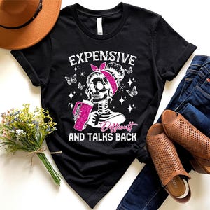 May include: Black t-shirt with a white graphic of a skeleton woman with pink hair and a pink headband holding a pink cup. The text "Expensive, Difficult and Talks Back" is printed in white below the graphic.