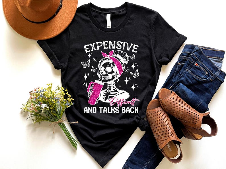 May include: Black t-shirt with a white graphic of a skeleton woman with pink hair and a pink headband holding a pink cup. The text "Expensive, Difficult and Talks Back" is printed in white below the graphic.