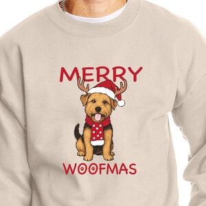 May include: A beige sweatshirt with the words "MERRY WOOFMAS" in red. The graphic features a dog wearing a Santa hat with antlers and a red scarf with white polka dots. A festive holiday design.