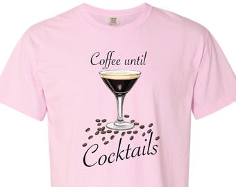 May include: A light pink t-shirt with the text "Coffee until Cocktails" in a script font. The shirt features an illustration of a coffee cocktail in a martini glass, surrounded by coffee beans.
