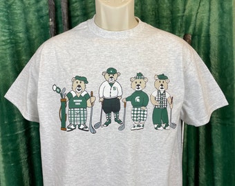 Puede incluir: Una camiseta gris con un gráfico de cuatro osos de dibujos animados vestidos con atuendos de golf y sosteniendo palos de golf. Los osos llevan pantalones de cuadros verdes y blancos, camisas a rayas verdes y blancas y sombreros verdes. El texto "Michigan State" está en la camisa de uno de los osos.