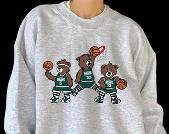 Puede incluir: Sudadera gris claro con un diseño de baloncesto. El gráfico muestra tres osos marrones con uniformes de baloncesto verdes y blancos, cada uno sosteniendo una pelota. Los osos están numerados 5, 33 y 12.