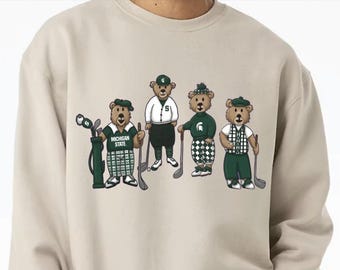 Puede incluir: Una sudadera beige de cuello redondo con un estampado gráfico de cuatro osos de peluche marrones vestidos con ropa de golf. Los osos llevan ropa verde y blanca, incluyendo gorros y jerséis, y sostienen palos de golf. Un oso está junto a una bolsa.